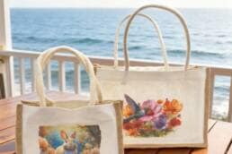 Deux sacs en jute personnalisés, grand et petit modèle, posés sur une table en bois avec vue sur la mer.